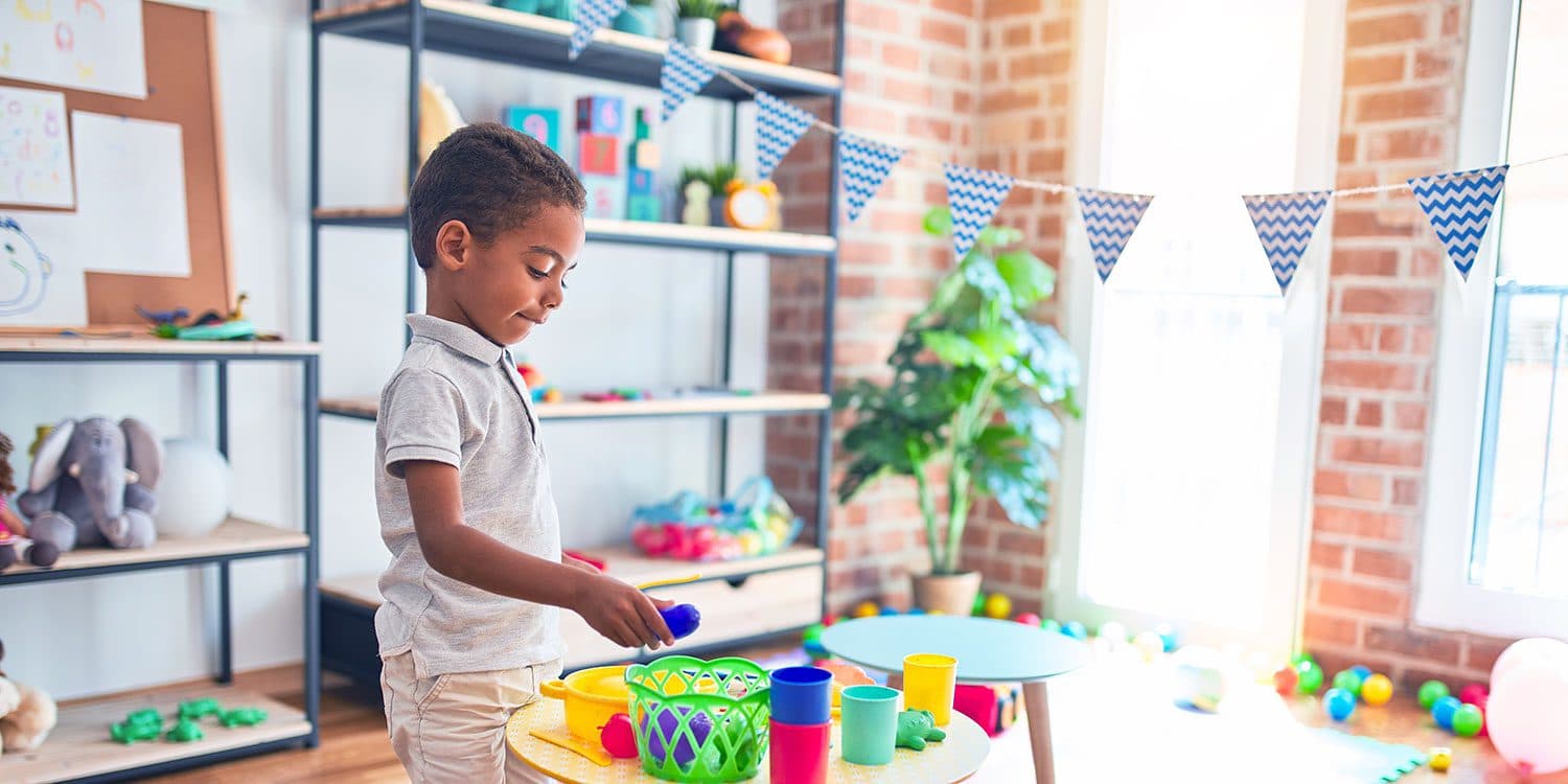 A boy playing with toy food and cups at a small table in a sunlit playroom