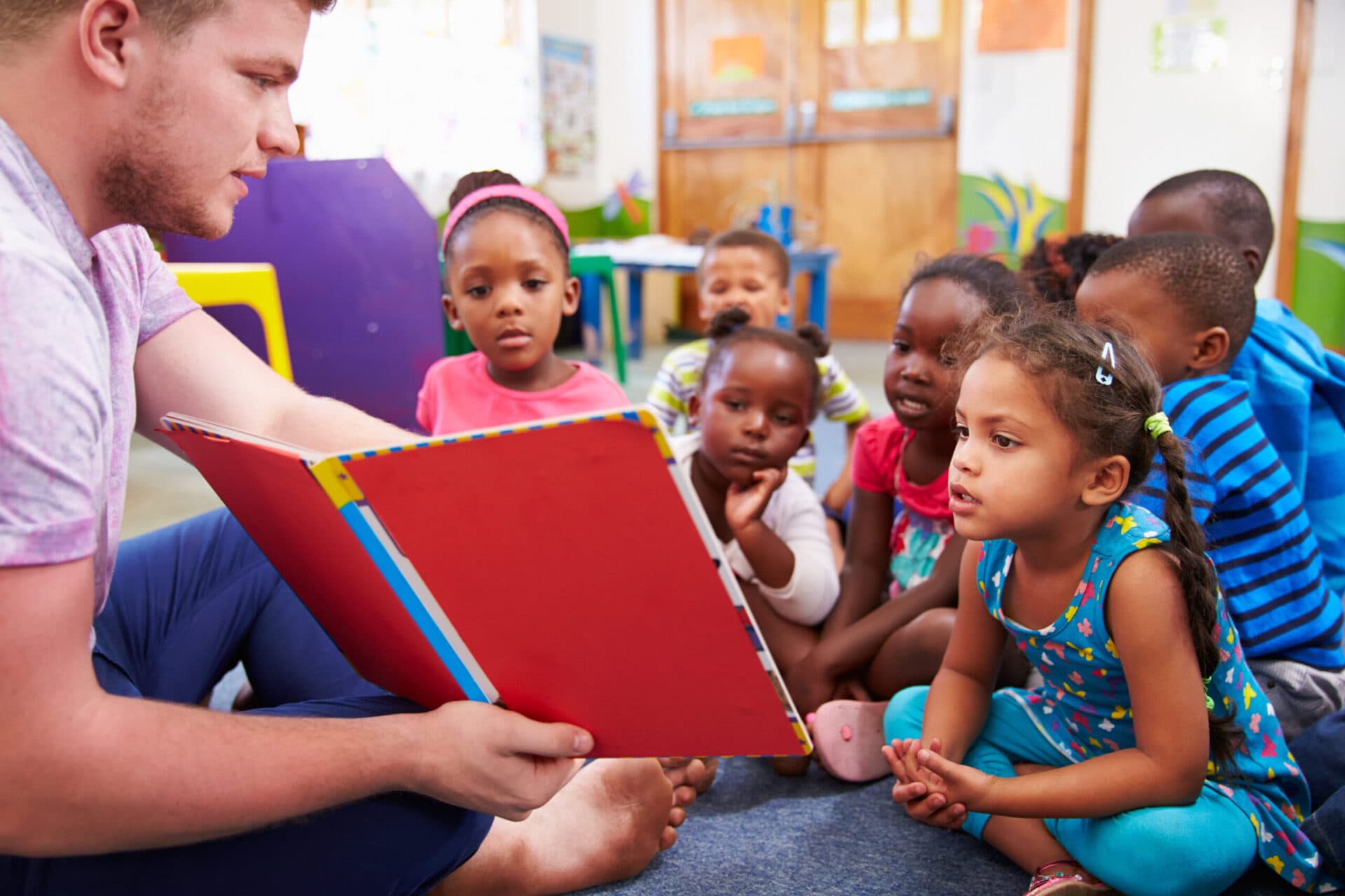 A teacher reading a large red picture book to a group of children seated on the floor