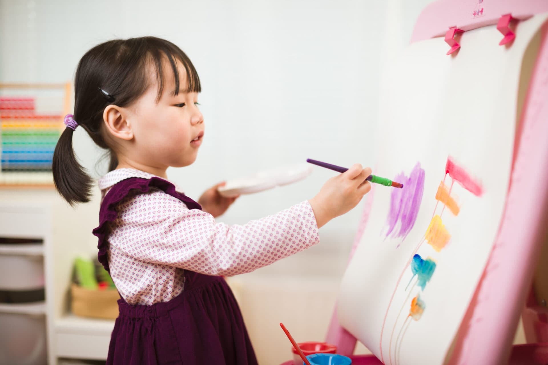 A girl in a pinafore painting colourful shapes on a large sheet of paper