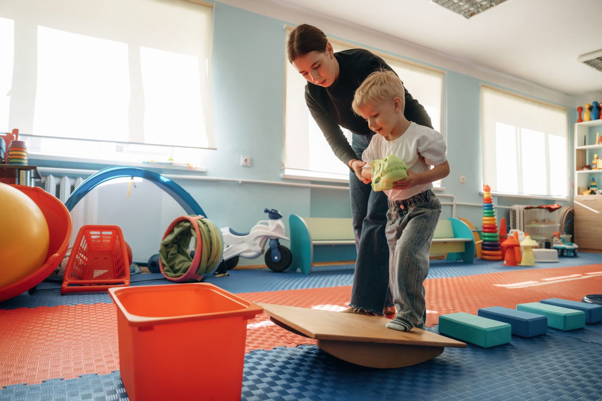 A teacher steadying a boy as he balances along a wooden beam in the gross-motor room