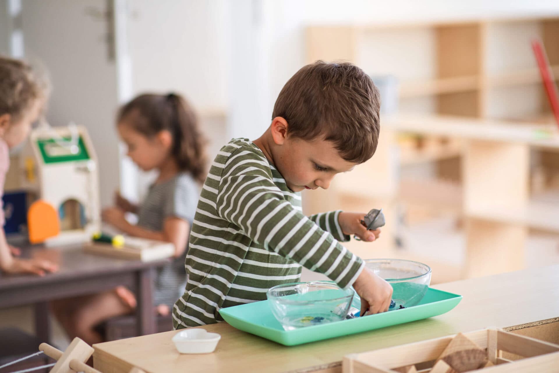A boy pressing his hands into a tray of green sensory material beside a classmate