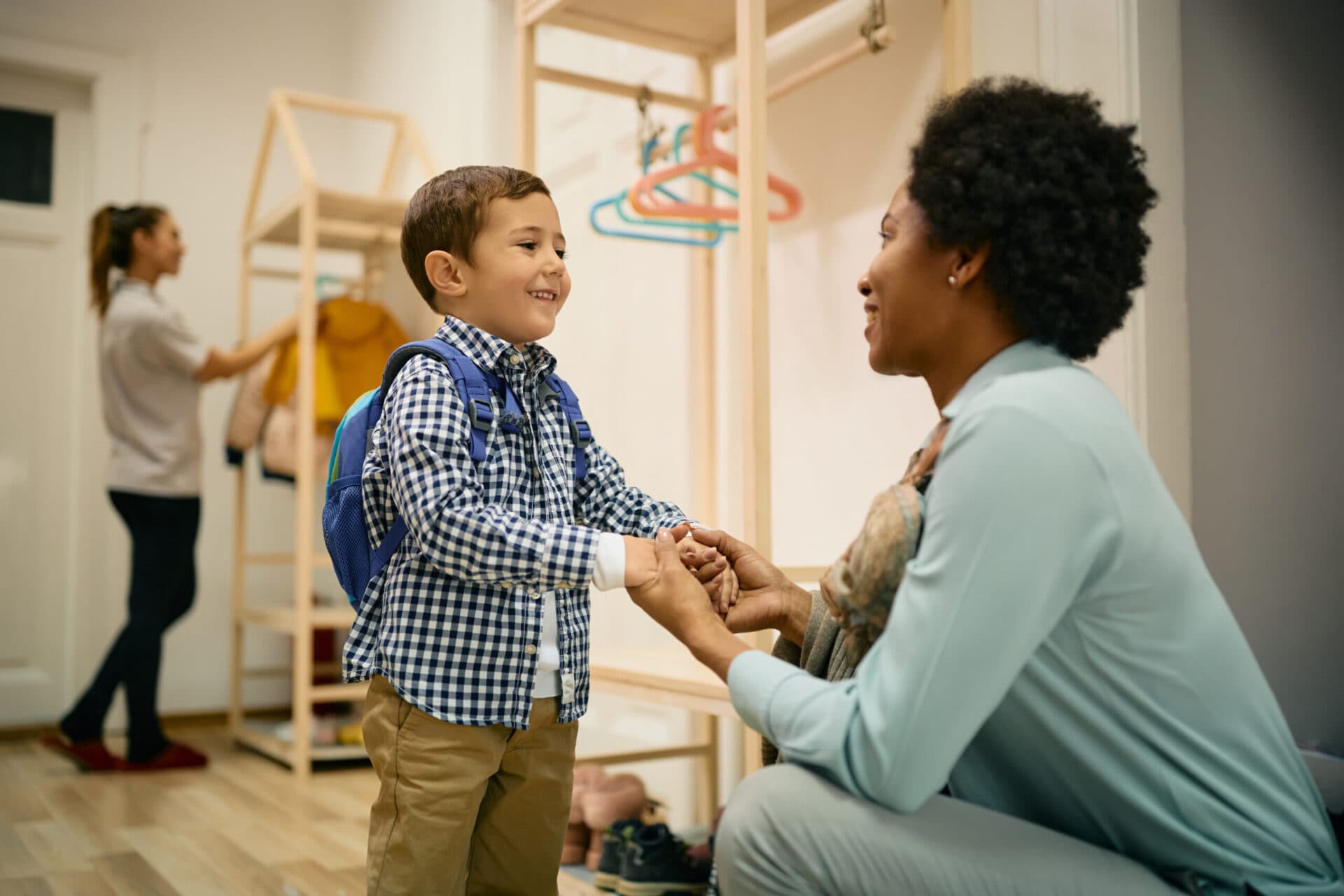 A teacher kneeling to greet a boy with a backpack at drop-off