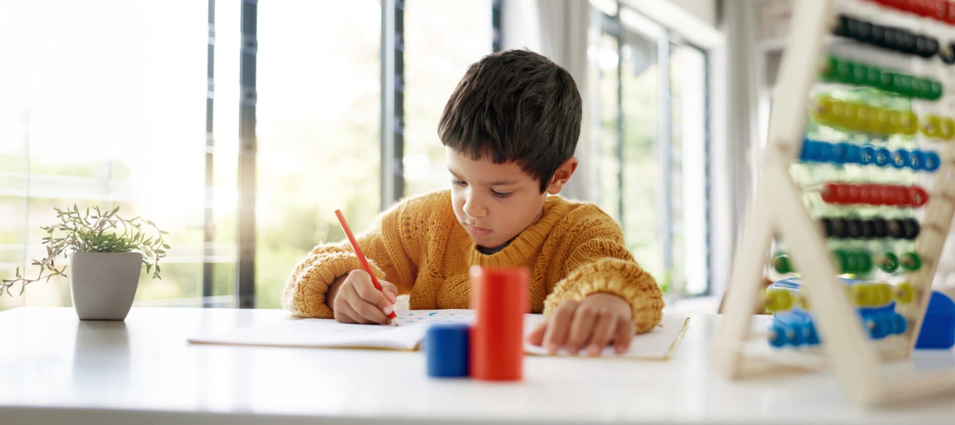 A boy in a mustard jumper drawing at a sunlit table with coloured pencils