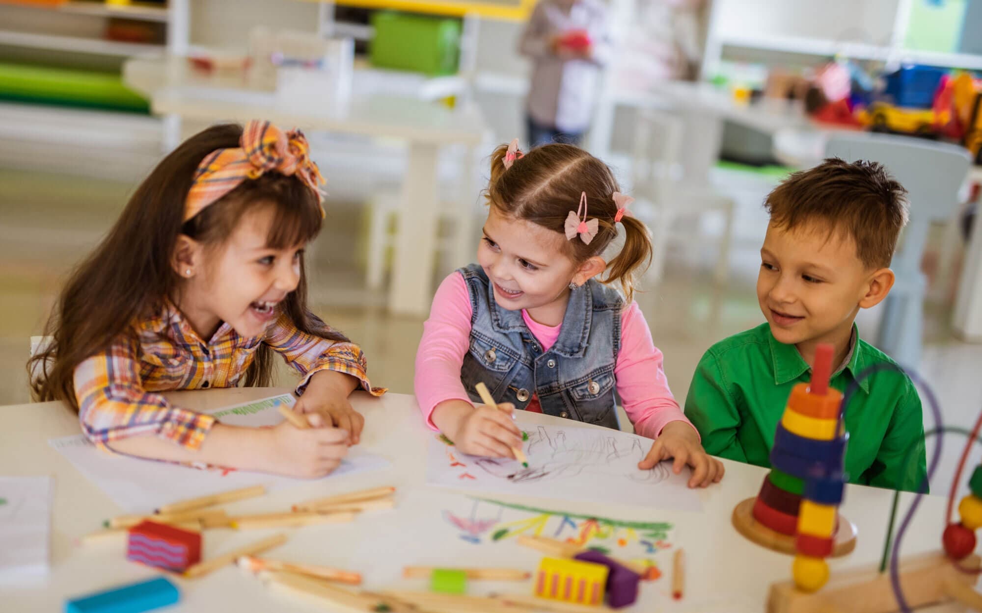 Three children drawing together at a classroom table with wooden toys nearby