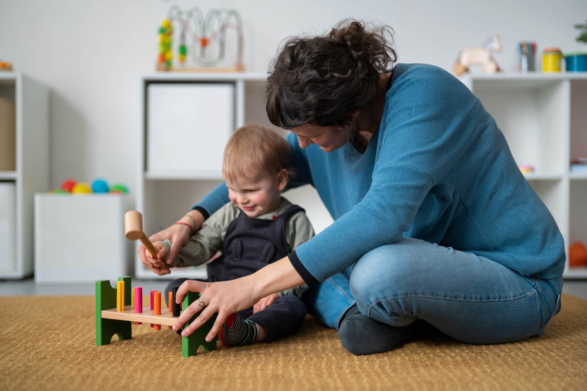 A teacher and toddler playing with a wooden hammer bench on the classroom rug
