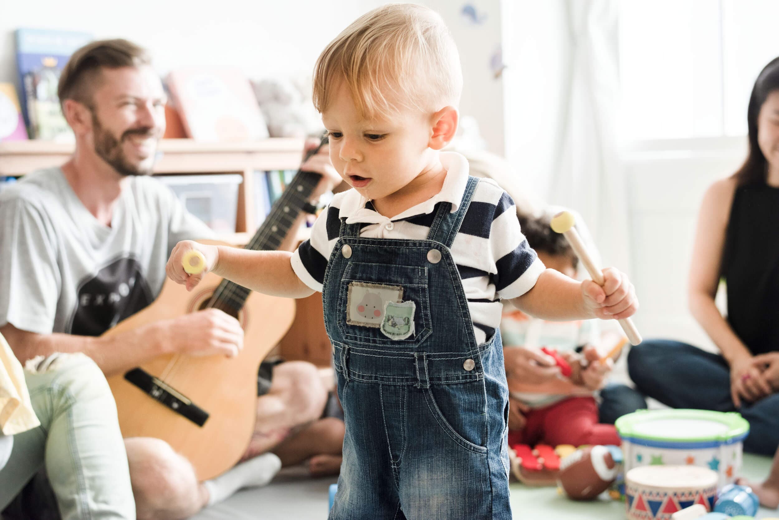 A toddler in dungarees exploring instruments while an adult plays guitar