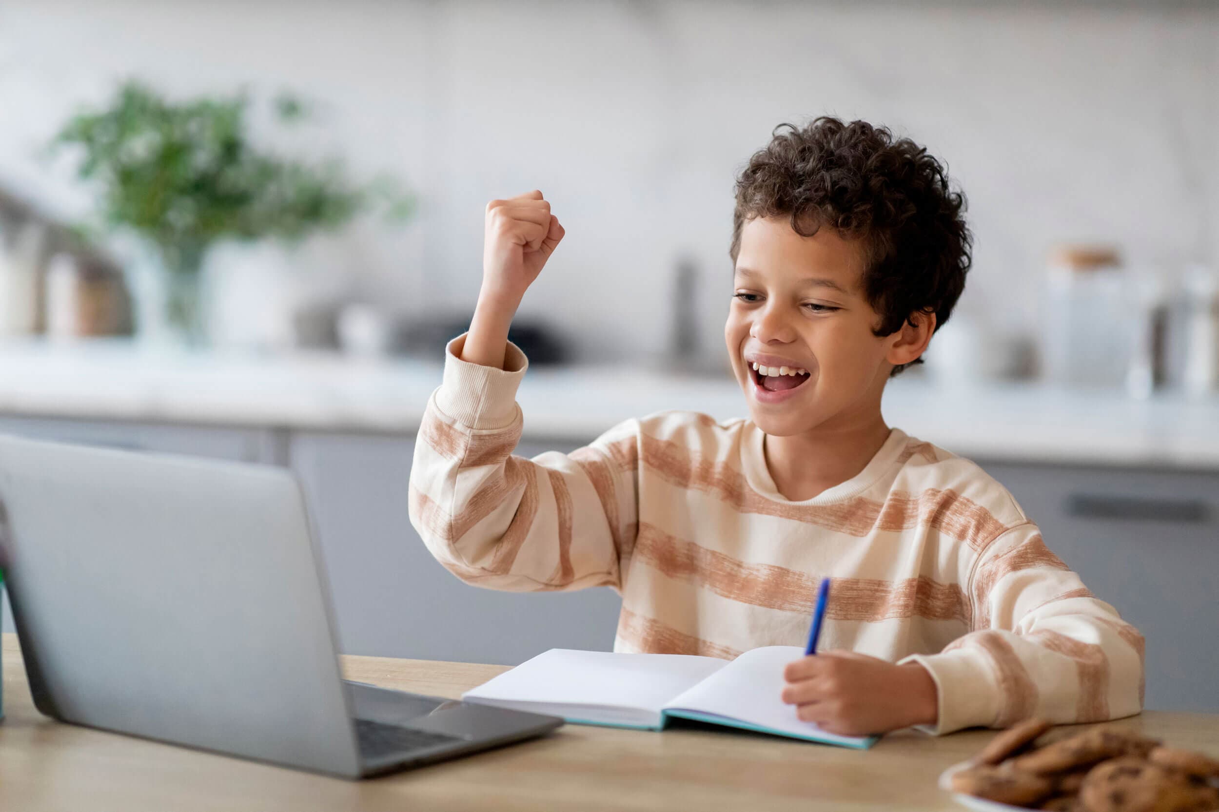 A boy at a laptop raising his fist in celebration with a notebook open