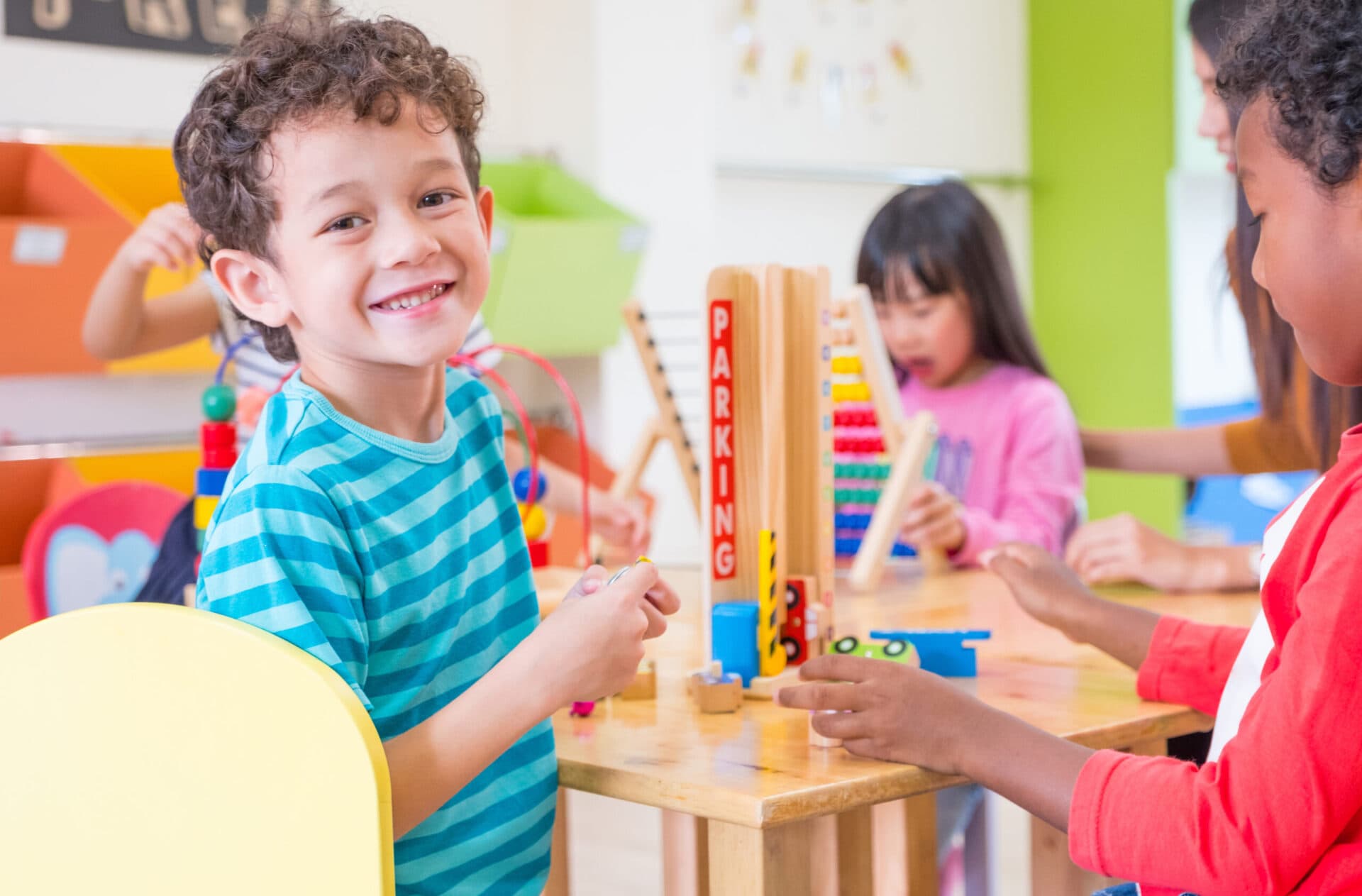 A smiling boy at a table of wooden toys with classmates and a teacher behind him