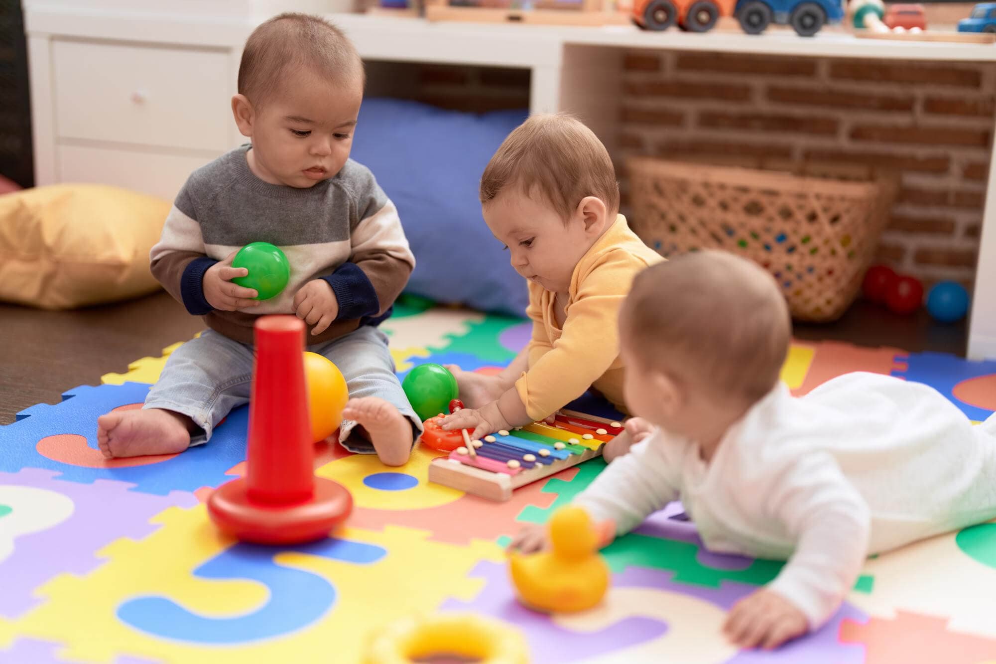Three babies playing with a ring stacker and xylophone on a colourful mat