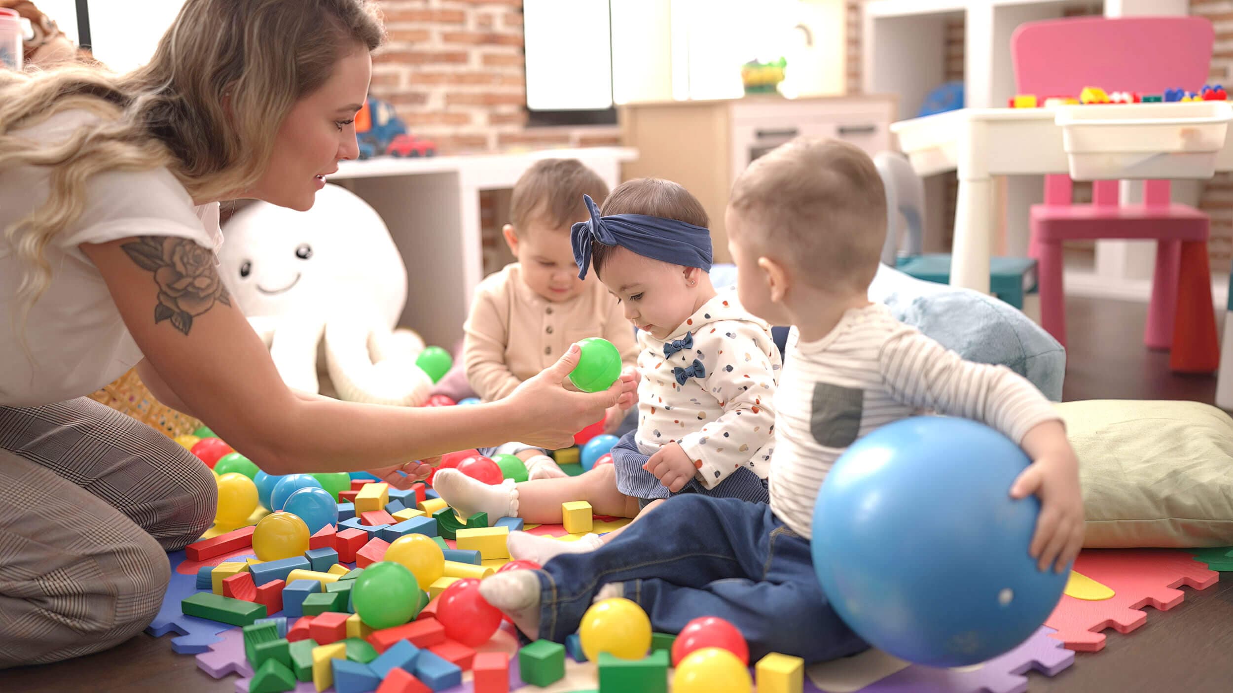 A teacher playing on the floor with toddlers among balls and soft toys